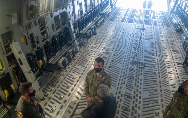 The image shows the interior of a large military cargo aircraft, likely a C-17 Globemaster III, with several military personnel and civilians gathered inside. The group appears to be engaged in a briefing or tour of the aircraft's cargo bay. All visible individuals are wearing face masks, suggesting this was taken during the COVID-19 pandemic period.