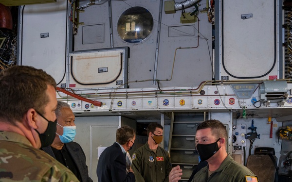 A group of five individuals are gathered inside what appears to be a military aircraft, likely a large transport or tanker aircraft. Two personnel in olive drab flight suits with U.S. flag patches are briefing or presenting to others, including a soldier in camouflage uniform and two civilians. All individuals are wearing face masks, suggesting this was taken during the COVID-19 pandemic period.
