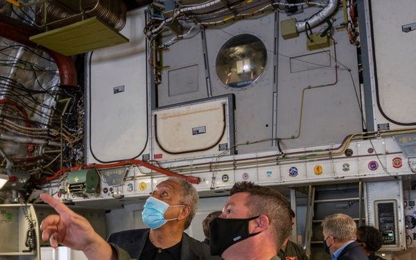 Two individuals wearing face masks are inside what appears to be a large military aircraft, likely a cargo or transport plane. A civilian in a dark jacket is pointing upward at the aircraft's interior systems while a military personnel member in an olive flight suit with a U.S. flag patch observes. Several other individuals are visible in the background, suggesting this is a guided tour or official visit.