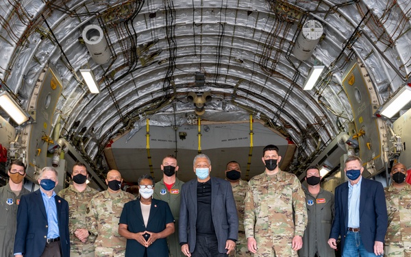 A group of approximately 13 individuals, including military personnel in camouflage uniforms and civilians in business attire, pose for a formal group photograph inside the cargo bay of a large military transport aircraft, likely a C-17 Globemaster III. Most individuals are wearing face masks, suggesting this photo was taken during the COVID-19 pandemic period. The group appears to represent a mix of military and civilian leadership.