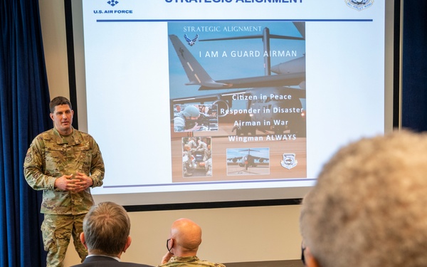 A U.S. Air Force military personnel in camouflage uniform is delivering a briefing or presentation to a small audience in a conference room. The presentation slide displayed behind the speaker is titled 'Strategic Alignment' and features the theme 'I Am a Guard Airman' with bullet points including 'Citizen In Peace, Responder in Disaster, Airman in War, Wingman ALWAYS.' The setting appears to be an official military briefing room with U.S. and Tennessee state flags visible.