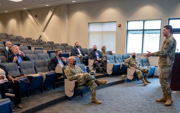 A military briefing or presentation is taking place in a small auditorium or conference theater. A uniformed service member stands at a podium addressing a small, socially distanced audience consisting of both military personnel in camouflage uniforms and civilians in business attire. A refreshment table with water bottles and snack bars is visible in the foreground.