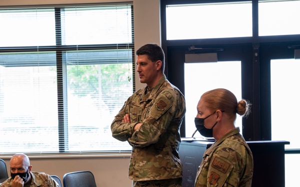 Three military personnel in OCP (Operational Camouflage Pattern) uniforms are gathered in what appears to be a briefing or conference room. Two individuals are standing near a podium while a third is seated in an auditorium-style chair. All three are wearing black face masks, suggesting this was taken during the COVID-19 pandemic period.
