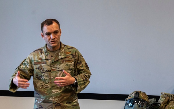A U.S. Air Force military officer in OCP (Operational Camouflage Pattern) uniform is presenting or briefing in what appears to be a conference or training room. Behind him is a table displaying various law enforcement and security equipment. A projection screen is visible on the wall behind him.