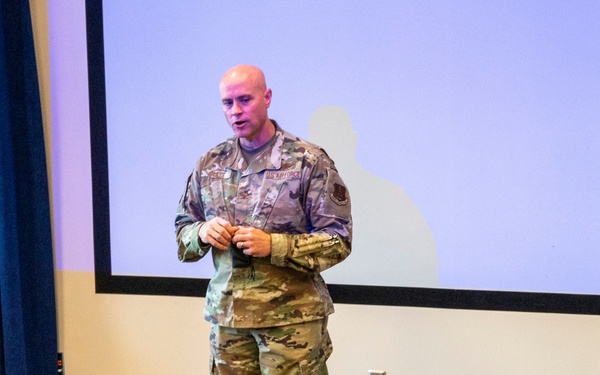 A U.S. Air Force service member in OCP (Operational Camouflage Pattern) uniform is delivering a briefing or presentation in a conference or auditorium setting. He is standing in front of a large projection screen with a silhouette image displayed. At least one audience member is visible from behind in the foreground.