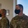 Two U.S. Army soldiers in OCP (Operational Camouflage Pattern) uniforms are standing indoors, both wearing black face masks. The soldier in the foreground is identified by his name tape as 'MELSON' and wears a Staff Sergeant rank insignia. Both soldiers appear to be at attention or observing something off-camera in what appears to be a military facility.