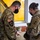 Two U.S. Army soldiers in OCP (Operational Camouflage Pattern) uniforms and black face masks are reviewing documents or a notebook at a table inside a building. The soldier on the left displays a 2nd Infantry Division patch and U.S. flag on their sleeve, while the soldier on the right displays what appears to be a 4th Infantry Division patch. They appear to be in a briefing or administrative setting.