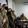 A group of military personnel wearing face masks are gathered in what appears to be a classroom or briefing room setting. Two individuals in U.S. Army multicam uniforms are visible on the left, alongside civilians and personnel in what appear to be Ukrainian military uniforms. The setting suggests a joint military training or coordination meeting.