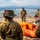 Joint Task Force-Bravo conducts overwater survival training A group of military personnel in camouflage uniforms are gathered on a sandy beach around an orange inflatable life raft. A civilian instructor in casual clothing appears to be demonstrating or explaining the raft's equipment and contents. The setting includes a large body of water with mountains visible in the background, suggesting a survival or water rescue training exercise.