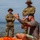 Joint Task Force-Bravo conducts overwater survival training Two soldiers in US Army OCP (Operational Camouflage Pattern) uniforms with boonie hats stand on a beach with arms crossed, observing a civilian man seated on the sand. The civilian, wearing a gray t-shirt and having visible tattoos on his arms, appears to be gesturing and holding a smartphone. The setting appears to be a coastal training or briefing environment, with mountains visible across the water in the background.