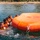 Joint Task Force-Bravo conducts overwater survival training A group of individuals wearing orange life vests are in the water alongside a large, inflated orange life raft. The scene appears to be a water survival training exercise taking place near a sandy beach with tropical vegetation. Additional personnel can be seen in the background near the shoreline.