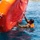 Joint Task Force-Bravo conducts overwater survival training A person in military-style clothing and an orange life vest is in open water, gripping and attempting to right or manage a large overturned orange inflatable life raft. The scene appears to be a water survival or rescue training exercise, likely conducted in a calm coastal or harbor environment. Additional swimmers are visible in the background, suggesting a group training scenario.