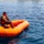 Joint Task Force-Bravo conducts overwater survival training A person in military camouflage uniform and an orange life vest is seated in an inflatable orange life raft on open water. Several swimmers are visible in the background, suggesting a water rescue training exercise or survival drill. The scene appears to take place in a calm coastal or lake environment under bright daylight conditions.