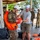 Joint Task Force-Bravo conducts overwater survival training A military training exercise is taking place at what appears to be a waterfront facility. A soldier in camouflage uniform and a white helmet labeled 'KONG' is being assisted by a civilian instructor in fitting an inflated orange life vest. Multiple other military personnel in camouflage uniforms are visible in the background, with additional orange life vests scattered on the wet concrete floor.