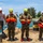 Joint Task Force-Bravo conducts overwater survival training Three military personnel in camouflage uniforms and safety helmets stand on a concrete pier or dock holding orange life vests, while a civilian instructor in casual clothing appears to be briefing or instructing them. The setting appears to be a tropical coastal location with turquoise water, trees, and industrial barrels visible in the background. This appears to be a water rescue or maritime safety training exercise.