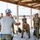 Joint Task Force-Bravo conducts overwater survival training A civilian instructor or briefer in a white t-shirt and baseball cap addresses a group of military personnel in camouflage uniforms under an open-sided shelter near a beach. The group appears to be receiving a briefing or training instruction, with personnel standing and one individual crouching on a small stool. The setting suggests a coastal military training exercise or pre-mission briefing.