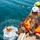 Joint Task Force-Bravo conducts overwater survival training Military or rescue personnel in camouflage uniforms and helmets are conducting a water survival or rescue training exercise from a pier or dock. One individual has just jumped into the water and is visible in the splash zone wearing an inflated orange life vest and green helmet. Several other swimmers are visible in the background water, suggesting a group training evolution.