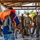 Joint Task Force-Bravo conducts overwater survival training A military training or briefing session is taking place under an open-air covered structure, likely in a tropical location. A civilian or instructor wearing a blue patterned outfit is demonstrating the use of an orange life vest/survival vest to a group of military personnel in camouflage uniforms. The setting and equipment suggest water survival or rescue training.