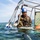 Joint Task Force-Bravo conducts overwater survival training A military or rescue training exercise is taking place in open water. A person wearing camouflage uniform, a blue helmet, and a harness is partially submerged within a metal frame structure, interacting with a swimmer wearing goggles. The scene appears to be a water survival or dunker/egress training exercise.
