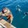 Joint Task Force-Bravo conducts overwater survival training A person in military camouflage uniform and an orange life vest is climbing down a ladder or ramp from a dock or vessel toward the water. Three individuals are already in the blue water below, wearing swim goggles and appearing to float or tread water. The scene appears to depict a water survival training or military swim qualification exercise.