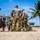 Joint Task Force-Bravo conducts overwater survival training A group of military personnel in camouflage uniforms are gathered on a sandy beach area, appearing to work on or examine something on the ground. The setting includes a tropical environment with palm trees, a concrete building, and a communications tower in the background. The group appears to be engaged in a training exercise or field operation.