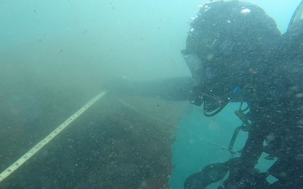 An underwater photograph showing a scuba diver conducting what appears to be an archaeological or scientific survey of a submerged structure or wreck. The diver is holding or placing a measuring scale/ruler against a flat, dark surface on the seafloor. The water visibility is moderate with suspended particulate matter visible throughout the frame.