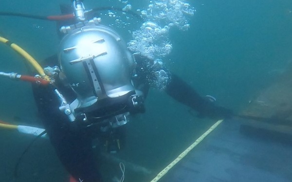 A commercial or military diver is working underwater alongside what appears to be the hull of a large vessel or submerged structure. The diver is equipped with a full-face helmet and yellow umbilical hoses, suggesting surface-supplied diving equipment. A measuring tool or ruler is visible against the hull surface, indicating an inspection or survey operation.