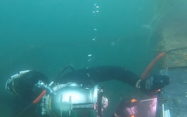An underwater diver is performing welding or cutting work on a submerged metal structure. The diver is equipped with a specialized helmet and full diving suit, with visible sparks/arc light at the work point. The image was taken underwater with characteristic green-tinted water visibility.