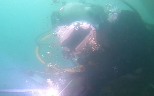 An underwater welder is performing wet welding operations on a submerged metal structure. A bright welding arc is clearly visible at the point of contact between the welding electrode and the metal surface. The diver is equipped with a specialized diving helmet and is working in murky, greenish-tinted water.