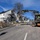 U.S. Army Corps of Engineers manages building renovation projects at the Baumholder Military Community A street construction or utility work site is underway in what appears to be a European residential area. A Volvo wheeled excavator (model EW150E) is actively operating on the road while workers in high-visibility gear supervise and assist. The scene is set on a clear sunny day with residential buildings and bare trees visible in the background.