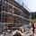 U.S. Army Corps of Engineers manages building renovation projects at the Baumholder Military Community A construction or renovation site showing a multi-story building covered in scaffolding, with exposed brick and stonework visible on the facade. A worker wearing an orange high-visibility vest and white hard hat is observing the site from the foreground. Construction materials including timber planks, sand, and debris are scattered at ground level.