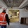 U.S. Army Corps of Engineers manages building renovation projects at the Baumholder Military Community An interior construction or renovation site is shown, with a worker in an orange high-visibility vest and white hard hat walking through the space. The ceiling is partially open, revealing metal framing, conduits, and exposed wiring. In the background, another worker is visible on a ladder performing overhead work.
