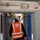 U.S. Army Corps of Engineers manages building renovation projects at the Baumholder Military Community A construction or safety inspector wearing an orange high-visibility vest labeled 'Europe District' and a white hard hat walks through a corridor undergoing renovation or construction work. The hallway shows exposed pipes, cables, and partially stripped ceiling elements, indicating active construction. The tiled walls and visible utility infrastructure suggest this is likely a commercial or institutional building under refurbishment.