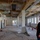U.S. Army Corps of Engineers manages building renovation projects at the Baumholder Military Community A construction or renovation site showing a large interior space with exposed ceiling infrastructure, dangling wires, and stripped surfaces. A worker wearing an orange high-visibility vest and white hard hat is observing the space from the right foreground. The room appears to be in an active gut-renovation phase with construction materials and tools scattered across the floor.