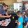 An Earth Day event at a home center store where participants are planting seedlings. A masked adult volunteer is demonstrating seed planting to children and adults using small pink pots and soil trays. A sign in the background advertises the 'Celebrate Earth Day' event scheduled for Saturday, April 24th from 12:00-14:00.