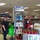 Two masked individuals stand at an Earth Day 2021 promotional display inside a retail store, likely in Guam based on the 'Hafa Adai! Guam Favorites' signage. The display features potted plants, reusable green bags, cleaning products, and an Earth Day poster decorated with colorful handprints. The setup promotes eco-friendly and sustainable products.