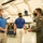 Three individuals are conversing inside what appears to be an aviation museum or aircraft hangar. Two men wearing matching blue polo shirts with an 'Aviation' logo are speaking with a person in an olive green military flight suit bearing a U.S. flag patch. All three are wearing face masks, suggesting this was taken during the COVID-19 pandemic period.