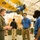 Three individuals wearing face masks are engaged in conversation inside what appears to be a military aviation museum. A person in an olive green flight suit with military patches is speaking with two individuals wearing matching blue polo shirts. A vintage U.S. Army biplane painted in yellow and blue is suspended from the ceiling in the background.