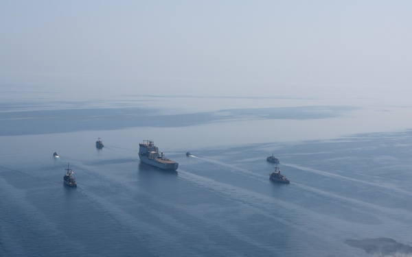 An aerial photograph showing a naval formation of multiple vessels underway on calm, open water under hazy atmospheric conditions. A large central vessel, likely a military or auxiliary ship, is flanked by numerous smaller escort or patrol craft arranged in a loose formation. A helicopter is visible in the lower right portion of the image, suggesting active aerial coordination.