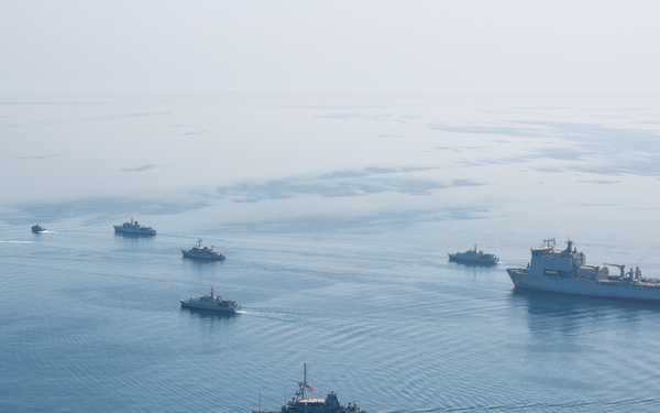 An aerial photograph shows a naval formation of approximately 10-12 military vessels underway on calm, open water under hazy atmospheric conditions. The formation includes one large vessel, likely a naval auxiliary or amphibious ship, accompanied by numerous smaller warships such as patrol craft or mine countermeasure vessels. The arrangement suggests a coordinated naval exercise or transit operation.
