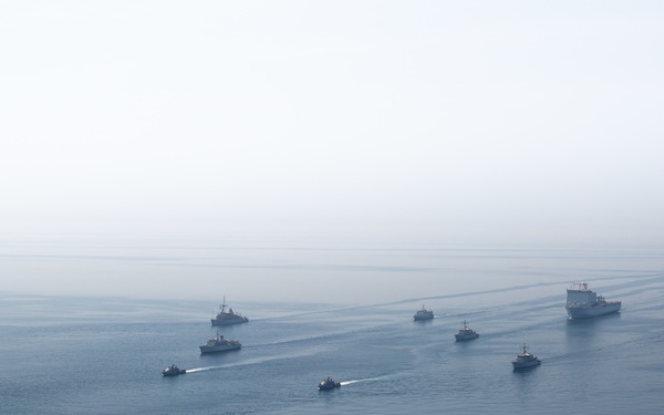 An aerial photograph captures a naval formation of approximately 10-12 military vessels sailing in a diagonal line across open ocean under overcast, hazy conditions. A military helicopter is visible on the left side of the frame, suggesting this is an active naval exercise or transit operation. The formation includes vessels of varying sizes, with at least one large amphibious or support ship prominent on the right side.