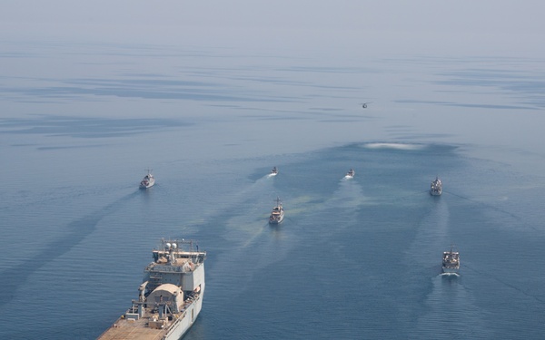 An aerial photograph shows a naval formation with a large vessel — appearing to be a catamaran-style military ship or expeditionary fast transport — leading a group of smaller patrol or coastal vessels across calm open water. The formation is arranged in a spread pattern with the lead ship at center and smaller craft flanking on both sides. Wake patterns are clearly visible behind each vessel, indicating active movement.