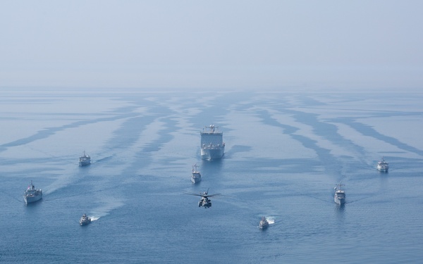 An aerial photograph captures a large naval formation of approximately 12 vessels sailing in a coordinated pattern across open blue water. A military helicopter flies low in the center foreground of the formation, while a large vessel — likely a command or amphibious ship — leads from the center rear. A circular water disturbance, likely caused by helicopter rotor wash, is visible in the lower foreground.