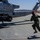 A U.S. Marine in flight suit and red helmet runs toward a Marine Corps attack helicopter on a desert airfield tarmac. The helicopter, marked 'MARINES' and identified as belonging to HMLA-269, is armed with what appear to be AGM-114 Hellfire missiles and rocket pods. The arid, desert terrain visible in the background suggests a location consistent with a Southwestern U.S. military installation.