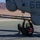 A military aviation technician in olive green flight suit is lying on his back on a tarmac, performing maintenance work on the underside of a military helicopter marked with the number '68'. The aircraft appears to be a gray military rotary-wing aircraft, likely a U.S. Marine Corps AH-1Z Viper or similar attack helicopter, positioned on a desert airfield. The technician is wearing a red and white flight helmet and appears to be working on a component resembling a gun barrel or structural rod mounted beneath the fuselage.