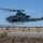 A U.S. Marines AH-1Z Viper attack helicopter is captured in low-level flight over a desert terrain, likely at or near a military airfield. The aircraft is in a gray camouflage scheme and is visibly armed with weapons systems on its stub wings. The arid, mountainous background and sparse desert vegetation suggest a southwestern U.S. location, possibly a training base.