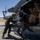 Two military personnel are working on a helicopter on a desert airfield tarmac. One crew member in a green flight suit is inspecting or loading a multi-barrel rotary cannon (likely an M134 minigun) mounted on the aircraft's door, while a second crew member in tan/desert camouflage sits inside the helicopter cabin. A second helicopter is visible in the background on the same tarmac.