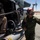 A U.S. Marine Corps crew member in a green flight suit and red helmet gives a thumbs-up signal beside a military helicopter on a desert airfield. The helicopter's door gunner position is visible with an M240 or similar belt-fed machine gun mounted, and ammunition belt loaded. The arid, desert terrain in the background suggests a location such as a Southwestern U.S. military installation.