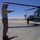 Military personnel are conducting ground operations around an attack helicopter on a desert airfield tarmac. A Marine in desert camouflage uniform is directing the aircraft with hand signals while other crew members are positioned nearby. The setting appears to be a arid, desert environment consistent with a southwestern U.S. military installation.