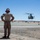 A military service member in desert camouflage (MARPAT) uniform stands on a desert airstrip observing a helicopter approaching or departing. The individual is wearing hearing protection with what appears to be an American flag design and has hands on hips in a supervisory stance. The arid, flat desert landscape and clear blue sky suggest a location such as a Southwestern U.S. military installation.